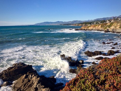 Waves crash against rocky shorelines under a clear blue sky, with mountains in the distance.