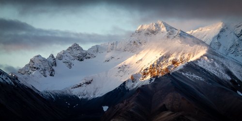 Snow-capped mountain peaks illuminated by sunlight, with dramatic clouds in the sky above.