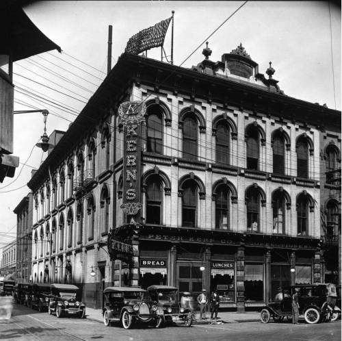 Historic black-and-white photo of a building with "Keen's" sign, vintage cars parked outside, and an American flag.