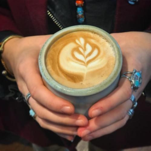A person holding a ceramic cup of latte art with a leaf design, adorned with multiple rings on their fingers.