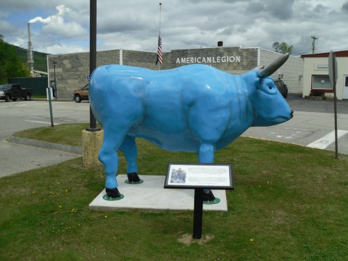 A large blue cow statue stands on a grassy area near a building, with an informational sign in front of it.