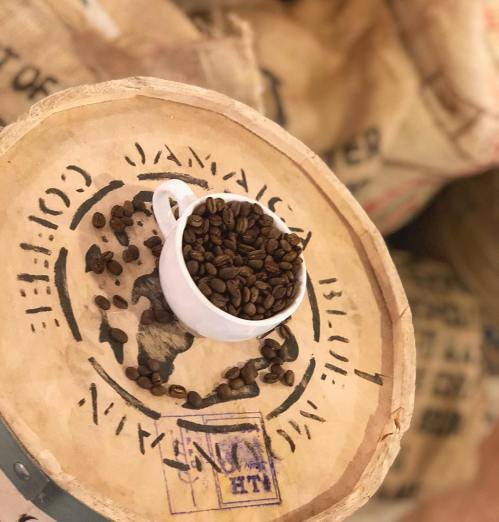 A small white cup filled with coffee beans sits on a wooden crate marked "Jamaica Coffee." Background features burlap sacks.