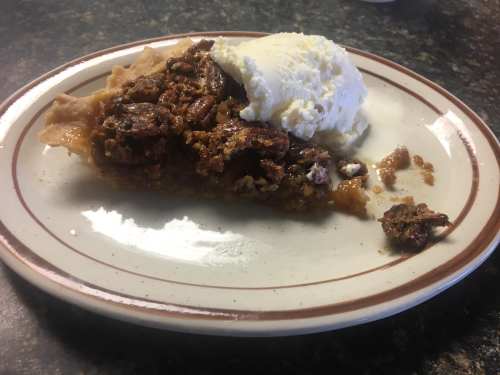 A slice of pecan pie topped with a scoop of vanilla ice cream on a white plate with a brown rim.