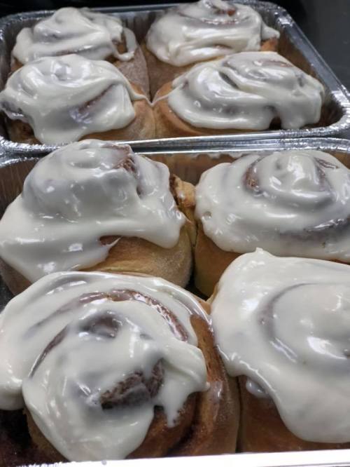 Freshly baked cinnamon rolls topped with creamy white icing, arranged in aluminum trays.