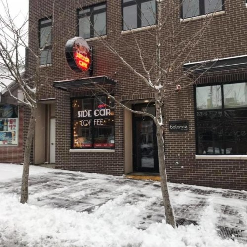 A cozy coffee shop entrance with a neon sign, surrounded by snow-covered trees and a snowy sidewalk.