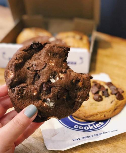 A hand holds a chocolate cookie with chunks, with a box of cookies in the background.