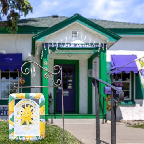A colorful "Welcome All" sign in front of a charming green and white building with a ramp and decorative awnings.