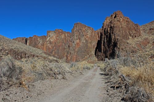 A dirt road leads through a rocky canyon with steep, colorful cliffs under a clear blue sky.