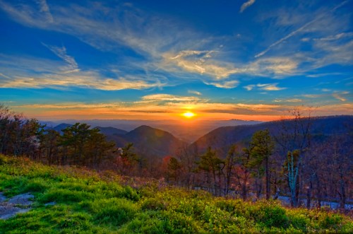 A vibrant sunset over rolling mountains, with colorful clouds and a foreground of green foliage.