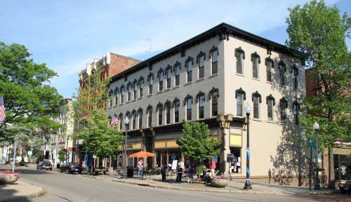 Historic building on a street corner, featuring large windows, trees, and outdoor seating, with cars parked nearby.