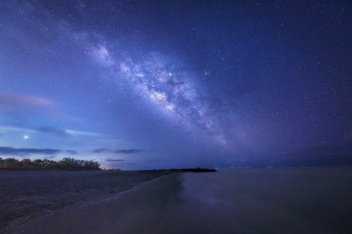 A serene beach scene under a starry night sky, featuring the Milky Way arching over calm waters.