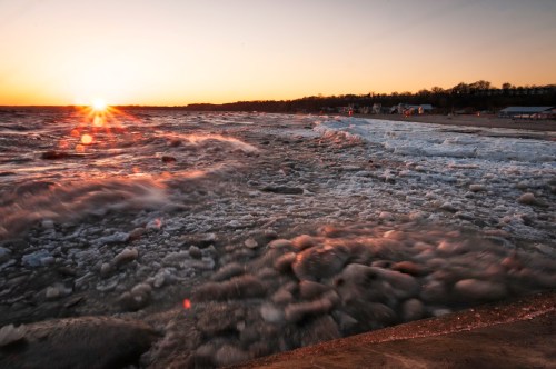 Sunset over a turbulent sea, with waves crashing and foam forming along the shoreline.