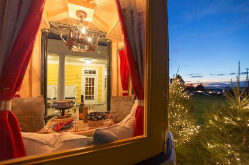 Cozy interior view of a cabin with a chandelier, wine, and snacks, framed by red curtains against a twilight sky.