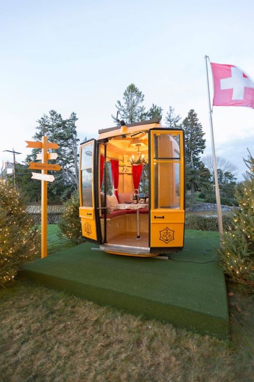 A cozy, yellow cabin with open doors, decorated with lights, set on green grass surrounded by trees and a Swiss flag.