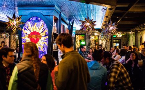 A lively bar scene with colorful decor, patrons socializing, and a vibrant stained glass feature in the background.