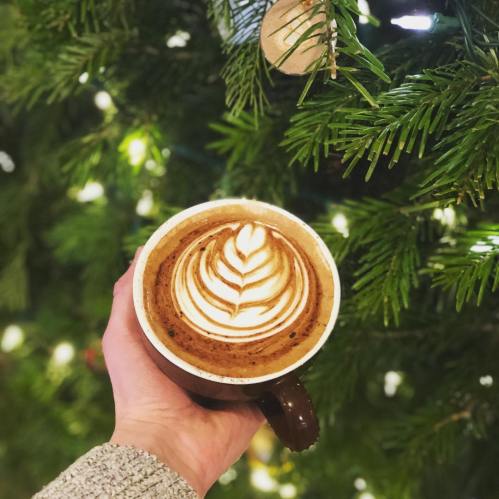 A hand holding a cup of coffee with latte art, set against a backdrop of a Christmas tree with lights.