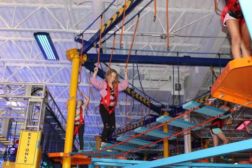 A girl balances on a high ropes course, wearing a harness and safety gear, with colorful structures and lights in the background.