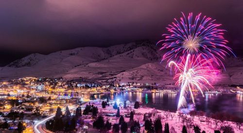 Colorful fireworks illuminate a snowy landscape and a lakeside town at night, with mountains in the background.