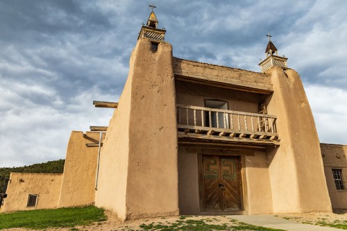 Adobe-style building with a wooden balcony and two spires, set against a cloudy sky.