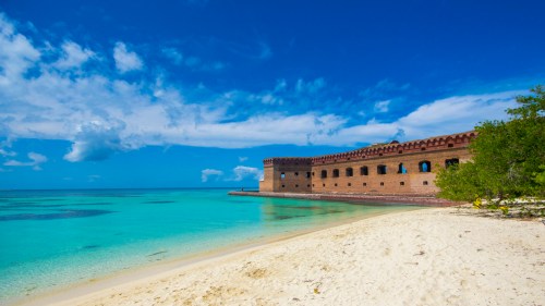A sandy beach with clear turquoise water and a historic brick fort under a bright blue sky with scattered clouds.