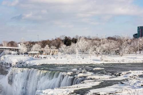 A winter scene of Niagara Falls, surrounded by snow-covered trees and a cloudy sky. People are visible near the edge.