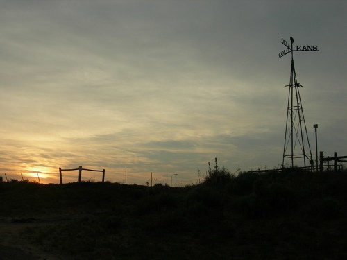 Silhouette of a windmill against a sunset sky, with a fence and grassy landscape in the foreground.