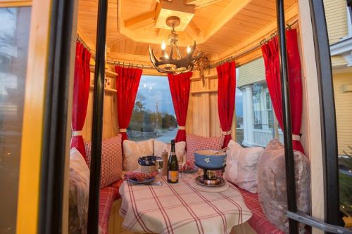 Cozy interior of a small cabin with red curtains, a chandelier, and a table set for a meal with champagne and a fondue pot.