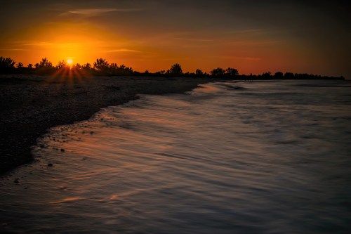 Sunset over a calm beach, with gentle waves lapping at the shore and silhouettes of trees in the background.