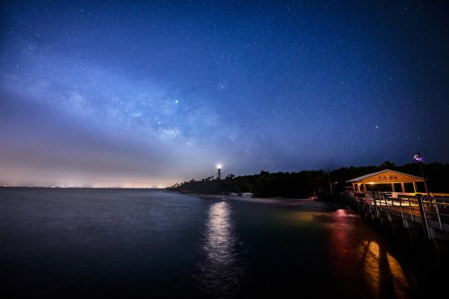 A serene night scene of a lighthouse by the water, under a starry sky with the Milky Way visible.