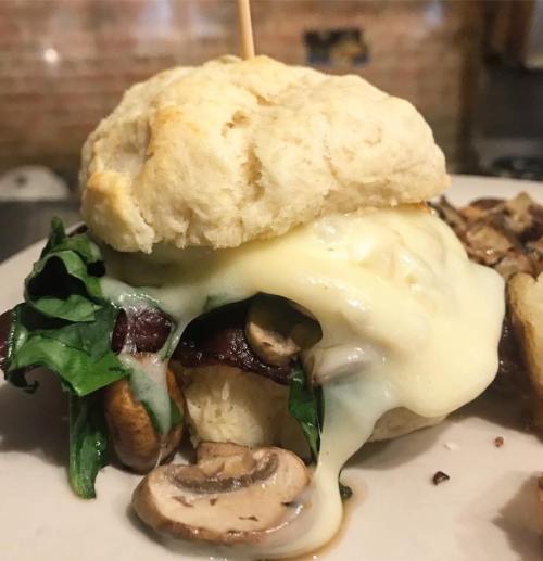 A close-up of a cheesy sandwich with mushrooms, spinach, and a biscuit-like bun, garnished with a toothpick.