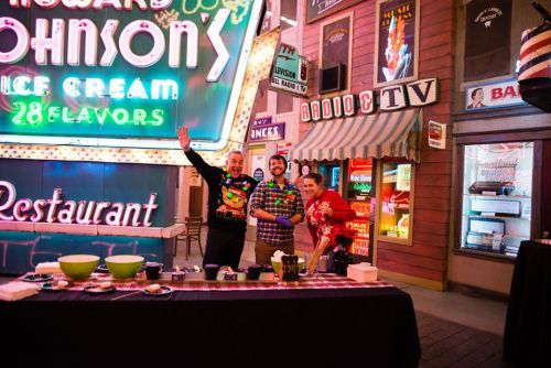 Three people in festive attire stand behind a table with bowls, smiling in front of colorful neon signs in a retro setting.