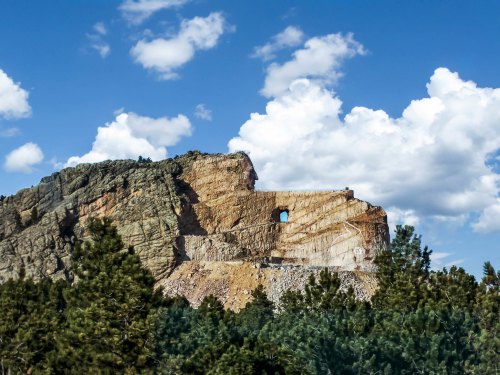 A rocky cliff with a distinct hole, surrounded by green trees and a blue sky filled with fluffy white clouds.