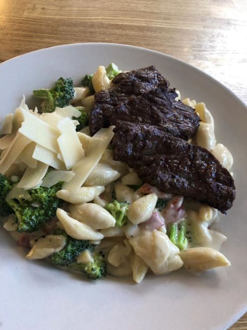 A plate of pasta with broccoli, creamy sauce, topped with slices of beef and shaved Parmesan cheese.