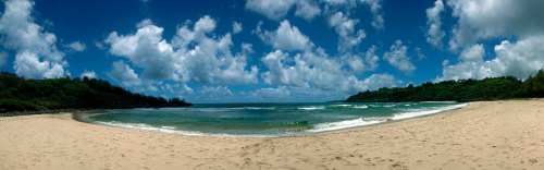 A panoramic view of a sandy beach with gentle waves, lush green hills, and a bright blue sky filled with clouds.