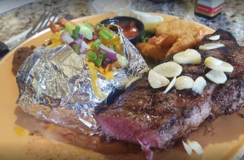 A plate featuring a grilled steak, a foil-wrapped baked potato with cheese, and a side of fried shrimp.