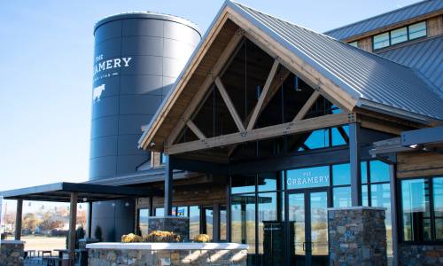 Exterior of a modern creamery building with a large silo, wooden beams, and glass entrance on a clear day.