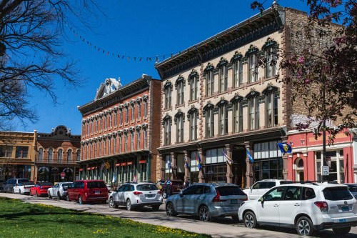 Historic buildings line a street with parked cars, under a clear blue sky and decorative string lights above.