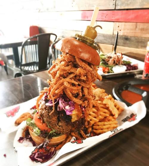 A towering burger topped with crispy onion rings, lettuce, tomato, and a pickle, served with fries on the side.