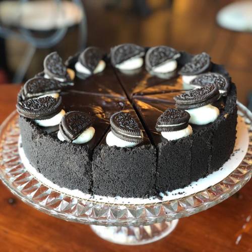 A round chocolate cake with Oreo cookies and cream topping, displayed on a glass cake stand. Slices are cut.
