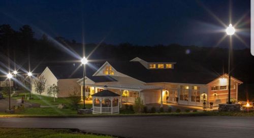 A well-lit building at night, featuring large windows and a gazebo, surrounded by landscaped grounds.