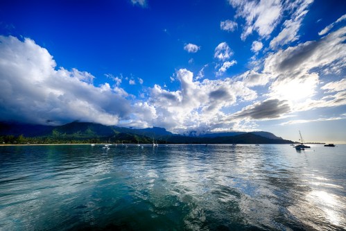 A serene seascape with calm waters, mountains in the background, and a vibrant sky filled with clouds and sunlight.