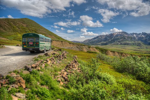 A green bus parked on a gravel road, surrounded by lush greenery and mountains under a partly cloudy sky.