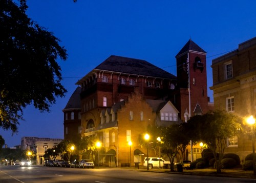 Historic brick building illuminated by streetlights at dusk, with trees lining the street and a clear blue sky.