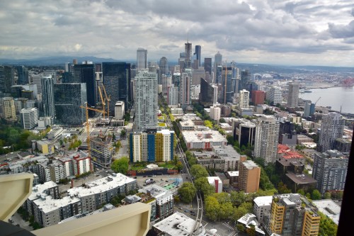 Aerial view of a city skyline with modern buildings, greenery, and a harbor under a cloudy sky.