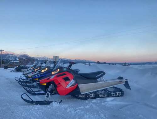 A row of snowmobiles parked on a snowy landscape with mountains in the background during sunset.