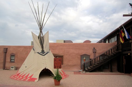 A traditional teepee stands in a courtyard, with a building and flags in the background under a cloudy sky.