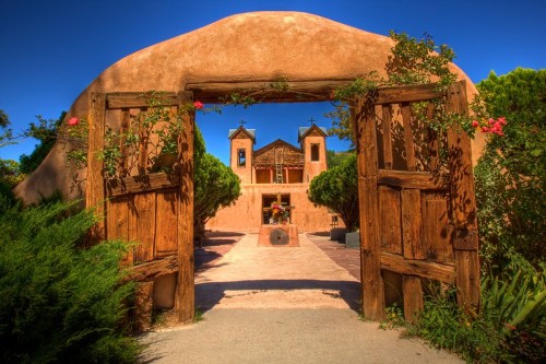 A rustic wooden gate opens to a serene courtyard with a church and vibrant greenery under a clear blue sky.