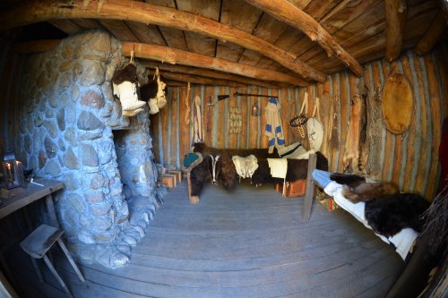 Interior of a rustic, ancient dwelling with stone walls, wooden beams, and fur garments hanging on the walls.
