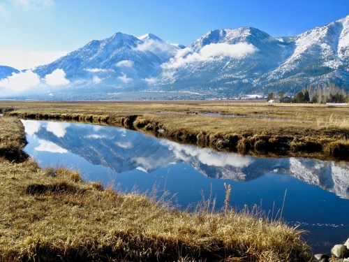 A serene landscape featuring a calm river reflecting snow-capped mountains under a clear blue sky.