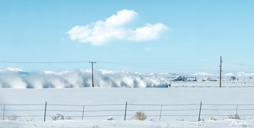 A snowy landscape with distant mountains, a cloud in the sky, and a fence line in the foreground.